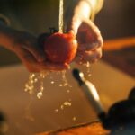 A person washing a fresh tomato under a faucet in warm kitchen lighting, highlighting cleanliness and health.