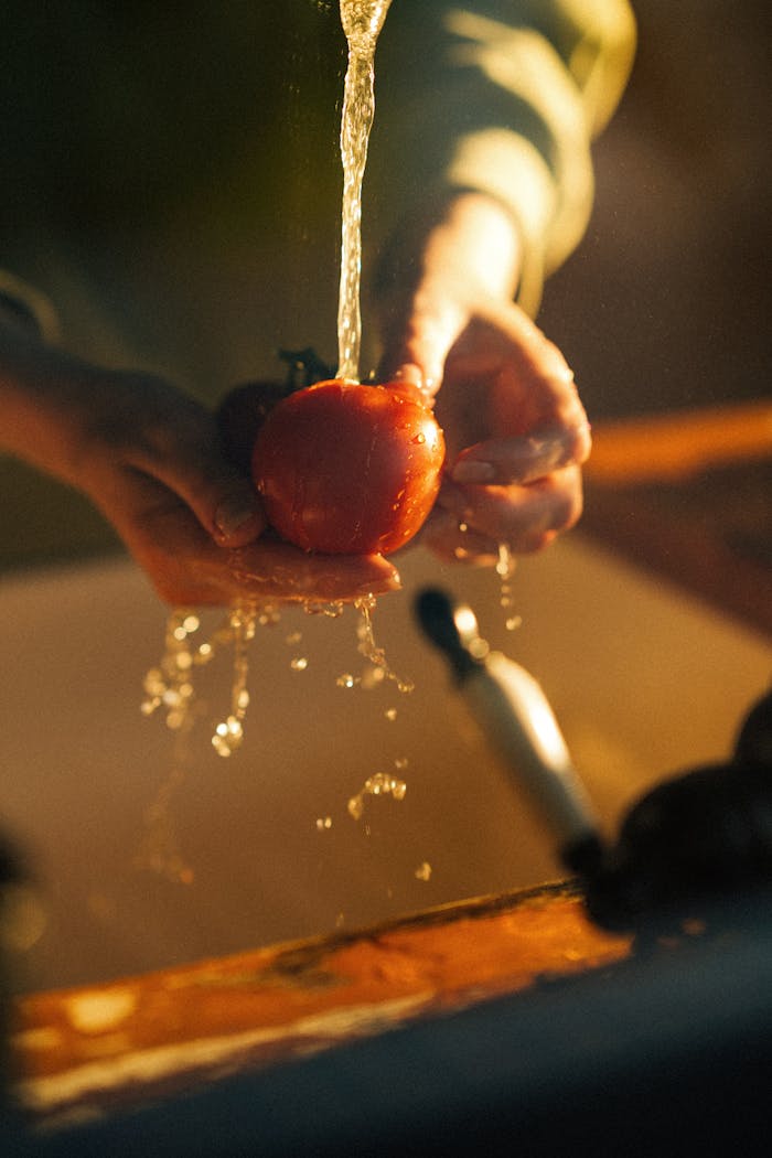 A person washing a fresh tomato under a faucet in warm kitchen lighting, highlighting cleanliness and health.