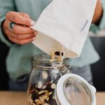 Woman pouring mixed nuts from a cotton bag into a glass jar, showcasing eco-friendly storage.