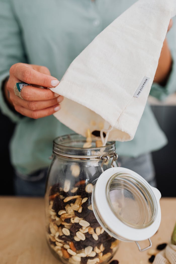 Woman pouring mixed nuts from a cotton bag into a glass jar, showcasing eco-friendly storage.